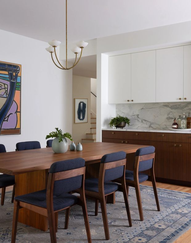 A refined dining and kitchen interior with a long mid-century-inspired timber dining table surrounded by upholstered navy dining chairs. The room features white walls and cabinetry, a dramatic veined stone backsplash, dark timber lower cabinetry, patterned area rug, sculptural chandelier, framed artwork, and decorative ceramics. A staircase and hallway are visible beyond the dining area.