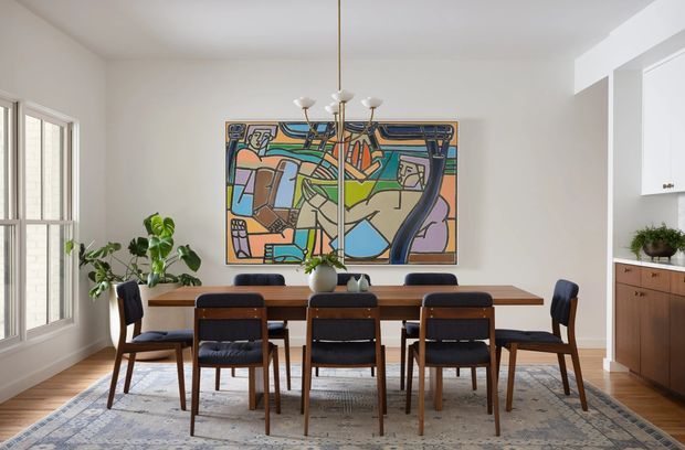 Bright, refined dining room with a long timber dining table surrounded by six dark upholstered mid-century-style chairs. A large colourful abstract artwork anchors the white wall, while potted plants, a patterned area rug, timber flooring, and a low timber sideboard add warmth. Tall windows provide abundant natural light, and a sculptural multi-arm pendant hangs centrally over the table.