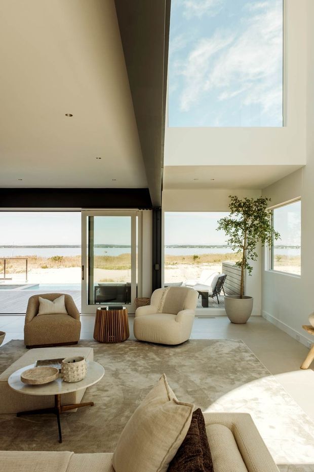 A bright, spacious coastal living room with large floor-to-ceiling glazing overlooking a sandy waterfront landscape. The room features a double-height ceiling with a large skylight, pale walls, light flooring, neutral upholstered seating, a textured area rug, a round coffee table, and a potted indoor tree. A dark exposed structural beam creates contrast across the ceiling, while the open glazing connects the interior to an outdoor pool or patio area.