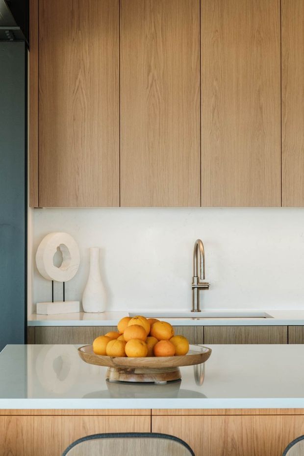A refined contemporary kitchen centered on a large island. Full-height light timber cabinetry with vertical slab doors spans the upper wall, paired with a pale stone or solid-surface splashback and benchtop. A brushed metal gooseneck tap and integrated sink sit beneath the cabinetry. The foreground island has matching timber joinery and a pale worktop, with curved dark-framed stools and a wooden bowl of citrus. A muted blue-grey wall and restrained sculptural accessories add contrast.