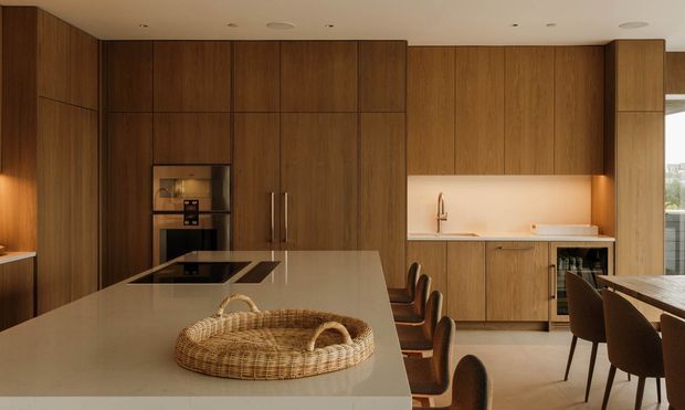 Warm contemporary open-plan kitchen and dining area dominated by full-height dark timber cabinetry. A large pale stone island with integrated cooktop occupies the foreground, while a timber-lined wall includes built-in appliances, concealed storage, sink, under-cabinet lighting, and wine storage. A dining table and upholstered chairs are visible to the right, with glazing beyond.