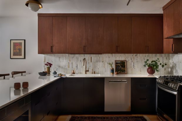 A warm, refined galley-style kitchen with dark lower cabinetry, extensive medium-dark timber overhead cabinetry, a white benchtop, and a vertically ribbed light backsplash. The kitchen includes a stainless-steel dishwasher, freestanding gas range, integrated sink, open shelving, and a small adjoining dining area with timber seating. Soft ambient lighting and restrained decor create a sophisticated mid-century-inspired atmosphere.