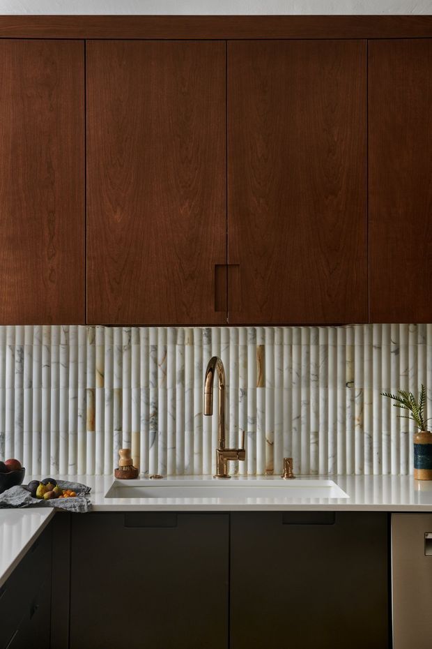 Close-up view of a refined kitchen sink area featuring dark walnut-toned flat-panel cabinetry, a white integrated benchtop and sink, a vertically ribbed light marble or stone splashback with subtle multicolour veining, and a polished brass gooseneck tap with matching fixtures. The composition is richly detailed and warmly lit, with a small fruit bowl and decorative objects on the counter.