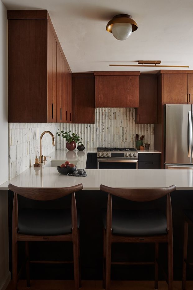A warm, richly detailed kitchen featuring dark timber cabinetry, a pale stone island with seating, a vertically textured light tile backsplash, stainless-steel appliances, and mid-century-inspired pendant and bar stools.