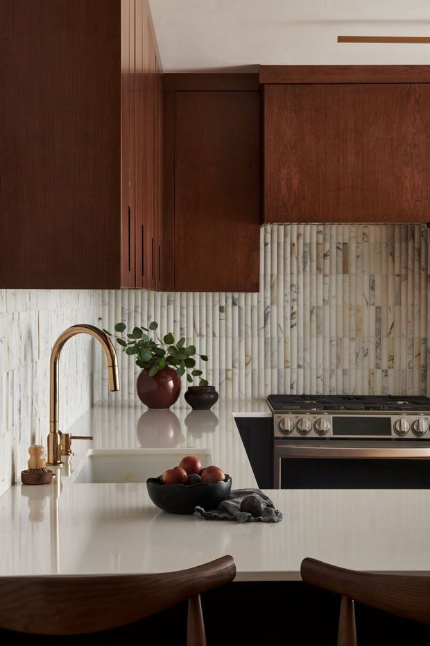 A warmly styled kitchen with dark walnut cabinetry, a pale vertically textured tile backsplash, a light stone island benchtop, and a stainless-steel range cooker. A curved brass-toned faucet and white undermount sink sit beneath the cabinetry, with dark wood dining chairs in the foreground and small decorative pottery and greenery on the counter.