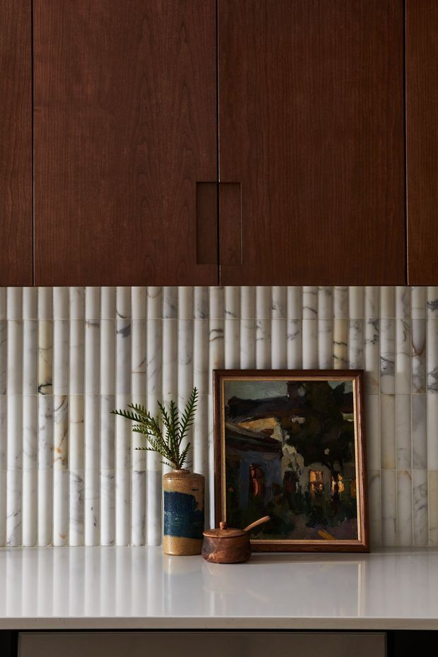 A close-up interior vignette featuring dark stained timber cabinetry with integrated handles above a white polished countertop. The backsplash is clad in vertically fluted, veined marble or stone tiles. A framed artwork, ceramic vase with foliage, and small wooden vessel sit on the counter, creating a warm, refined, richly textured composition.