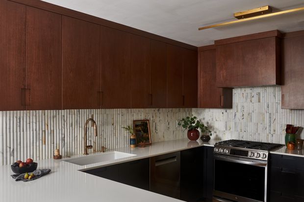 A refined galley-style kitchen with extensive dark timber overhead cabinetry, dark lower cabinets, a white stone-look benchtop, and a vertically ridged light backsplash. The kitchen includes a single-bowl undermount sink with a brushed metal tap, a freestanding stainless-steel range cooker, warm linear ceiling lighting, and a few decorative objects and plants. The overall composition is clean, warm, and strongly material-focused.