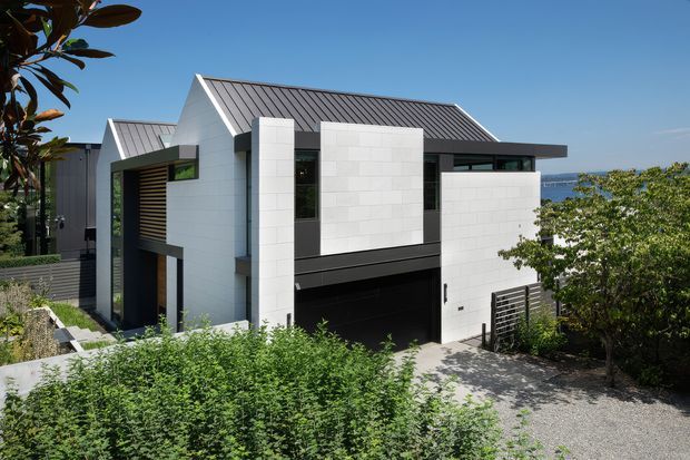 Exterior view of a contemporary multi-level residence on a sloping site, featuring crisp white masonry walls, dark metal standing-seam gabled roofs, deep charcoal window and door frames, horizontal timber screening, and an integrated double garage. Dense landscaping frames the foreground, with a partial distant water view beyond the house.