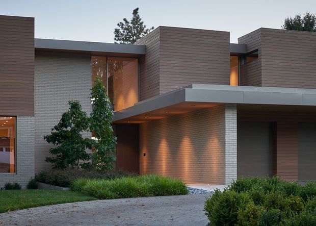 A contemporary multi-level residential facade photographed at dusk, featuring flat roof forms, projecting concrete-like canopies, warm horizontal timber-toned cladding, pale brick walls, recessed glazing, and integrated exterior lighting. Low landscaped planting and a paved approach frame the entrance and garage-like opening.