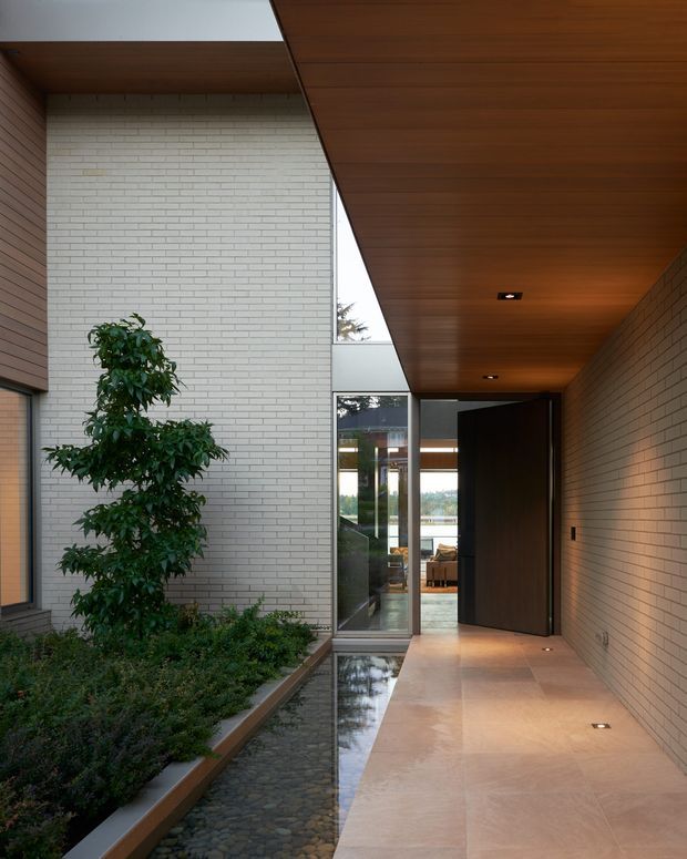 A sheltered contemporary entry courtyard and covered passage leading to a large dark timber door. Pale brick walls frame a narrow planted garden bed and pebble-lined reflective water channel, while warm timber soffits and large glazed panels create a calm, refined threshold.