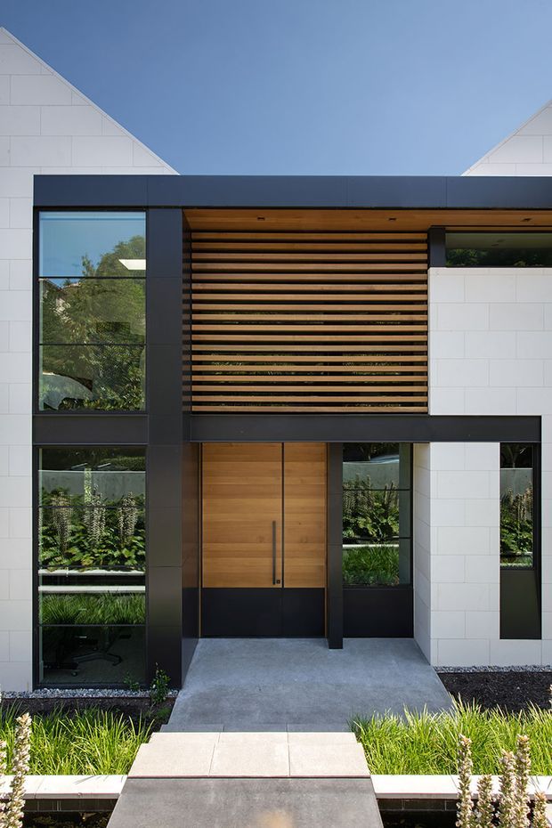 Front exterior view of a contemporary two-storey residence with a symmetrical gabled form, white rectangular masonry cladding, dark charcoal framing, large glazed openings, a timber entry door, and a horizontal timber privacy screen above the recessed entrance. A concrete path leads through landscaped planting to the front door.