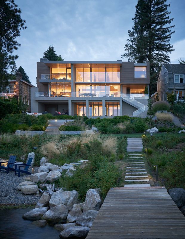 A contemporary multi-level house viewed from a landscaped waterfront garden. The home has broad floor-to-ceiling glazing, projecting horizontal roof and balcony planes, dark horizontal cladding, exposed concrete elements, and exterior stairs leading through layered planting to a timber boardwalk beside the water. Large boulders, ornamental grasses, mature trees, and outdoor seating create a natural, integrated landscape setting.