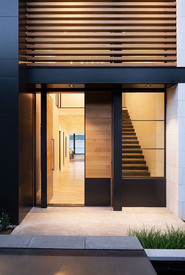 A contemporary residential entry facade viewed from outside, featuring a tall dark-framed pivot entry door with warm timber panels, a glazed stairwell, and a timber horizontal louvre screen above. The entry opens into a bright timber-lined hallway with a staircase visible behind glass. Pale paving, dark cladding, white masonry, and integrated warm lighting create a refined modern composition.