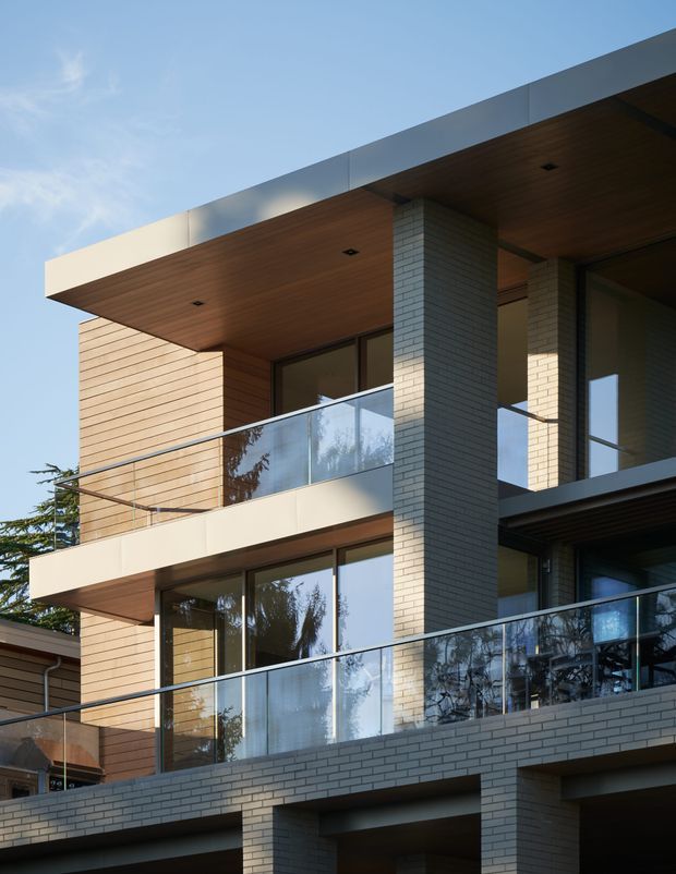 Exterior architectural photograph of a contemporary multi-storey residential building. The facade combines pale grey brick piers and walls, warm horizontal timber-clad sections, broad projecting flat roof slabs with timber soffits, large glazed openings, and glass balustrades on multiple balconies. The composition emphasizes strong horizontal lines, recessed balconies, and exposed geometric volumes against a clear sky.
