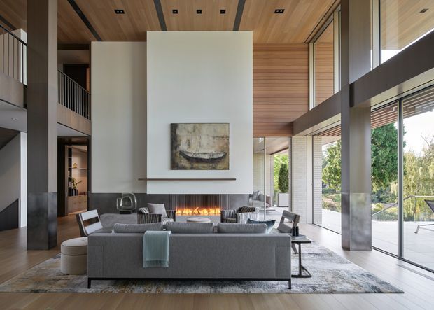 Double-height contemporary living room with a large grey sectional sofa arranged around a linear fireplace. The room features extensive floor-to-ceiling glazing, a timber-lined ceiling and upper wall panels, dark structural columns, polished concrete or stone fireplace surrounds, and a mezzanine level with a black balustrade. Neutral furnishings, a textured area rug, artwork, and views to landscaped grounds create a refined, warm interior.