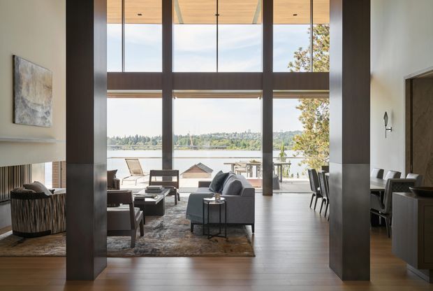 A refined, open-plan living and dining area with dramatic floor-to-ceiling glazing overlooking a waterfront landscape. The room features a high timber-lined ceiling, large dark structural columns, warm timber flooring, a contemporary fireplace, neutral upholstered seating, and a dining area set to the side. The expansive glazing and double-height volume create a bright, airy connection to the outdoors.