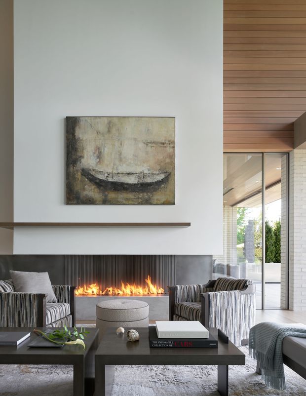 A refined contemporary living room centred on a long linear fireplace beneath a large white feature wall. The room includes two striped lounge chairs, a dark rectangular coffee table with books and small decor, a pale textured rug, and a glimpse of an adjacent glazed outdoor area. Warm timber ceiling and wall lining contrast with the white masonry and cool neutral furnishings.