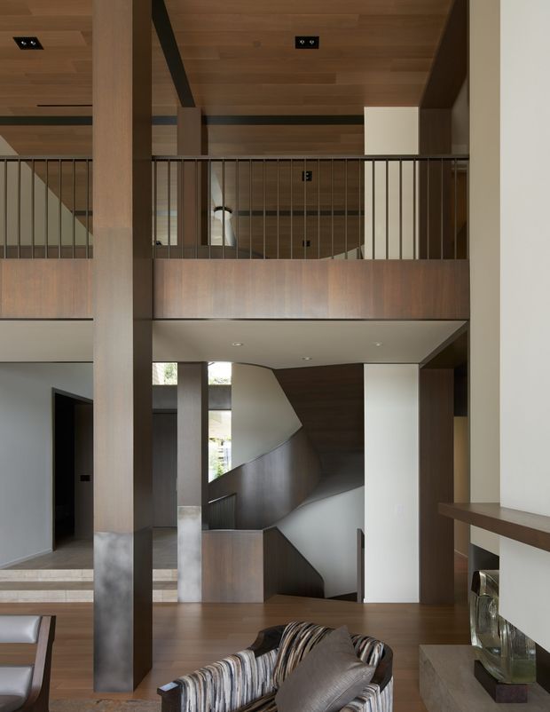 A sophisticated double-height interior focused on a sculptural stairway and mezzanine landing. Warm timber panelling and ceilings contrast with white plaster walls, dark structural beams, and slender black metal balustrades. A large timber-clad column, open void, built-in shelving, and partial living-room furnishings create a refined contemporary residential atmosphere.