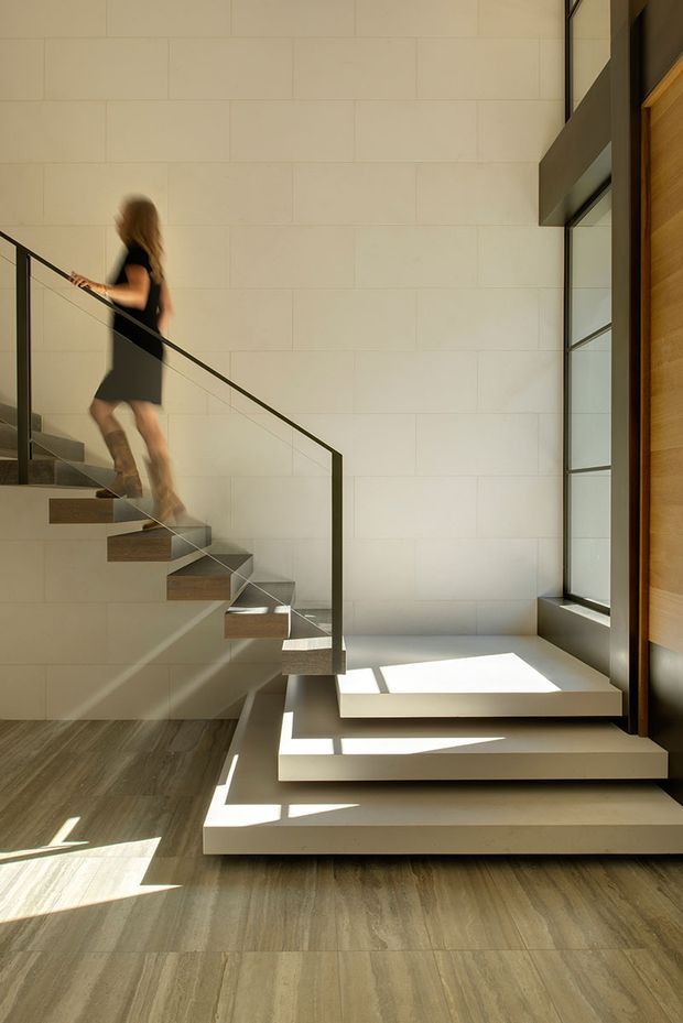 A sunlit contemporary stairway with floating timber-tread stairs, slim dark metal balustrades, and broad white tiled or stone-clad walls. Large vertical glazing and timber-framed detailing bring strong natural light into the double-height circulation space, creating sharp geometric shadows across the pale floor and steps. A blurred person is ascending the stairs.