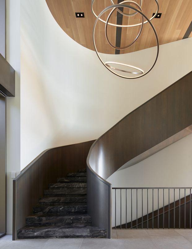 A dramatic interior stairway with a sweeping curved staircase, dark marble or stone treads, dark timber-panelled balustrade, and slim metal railing. The stairwell has tall white walls and a sculptural multi-ring pendant light beneath a curved timber ceiling feature, creating a refined contemporary architectural setting.