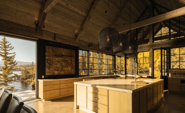 A spacious, warm kitchen in a rustic contemporary timber-framed home, featuring a large central island with pale stone benchtop, extensive timber cabinetry, black-framed glazed windows, exposed timber ceiling and beams, and oversized woven pendant lights. Autumn forest and a lake are visible through the glazing.