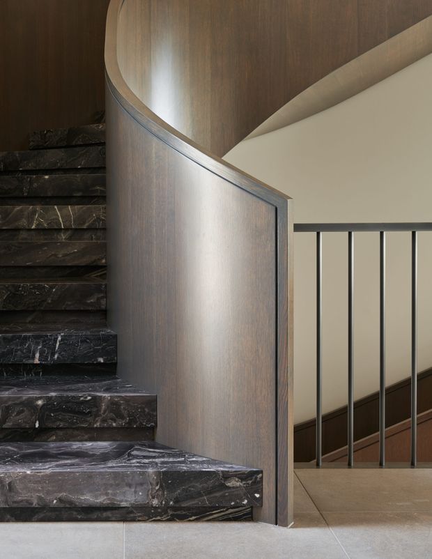 Close-up architectural photograph of an elegant stairway and landing. Dark, veined stone stair treads rise alongside a curved timber-clad balustrade or stair wall, with slim black metal railing and a contrasting light plaster wall. The lower floor appears to be large-format pale stone or tile. The composition emphasizes refined materials, curved joinery, and sculptural stair detailing.