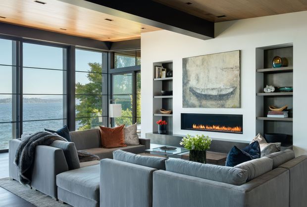 A contemporary living room with a large sectional sofa arranged around a linear built-in fireplace and wall-mounted artwork. Floor-to-ceiling dark-framed glazing opens views toward a body of water, while recessed shelving, a glass coffee table, and a warm timber-lined ceiling complete the space.