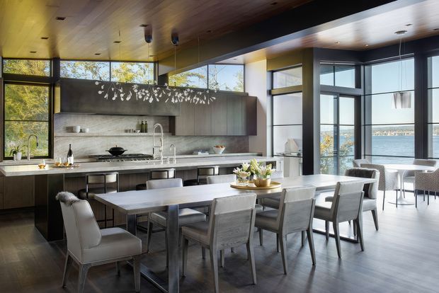 A sophisticated open-plan kitchen and dining area with dark timber cabinetry, a pale stone benchtop and backsplash, and a long dining table with upholstered chairs. Floor-to-ceiling glazing and expansive corner windows provide abundant natural light and views across the water. A timber-lined ceiling, black-framed windows, pendant lighting, and a decorative branch-like feature above the kitchen add visual interest.