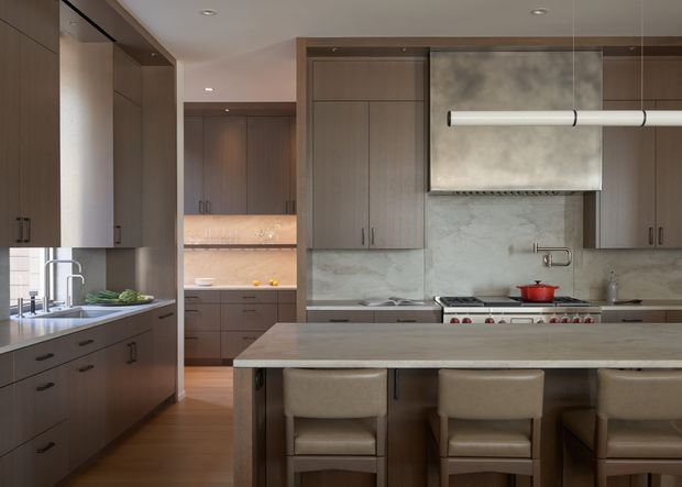 A refined contemporary kitchen with dark wood-toned cabinetry, a large pale stone island, integrated stainless-steel appliances, and three upholstered bar stools. The space includes a gas range with a sculptural metal rangehood, marble-look splashback, recessed ceiling lights, and a secondary preparation area visible through the opening at the rear.
