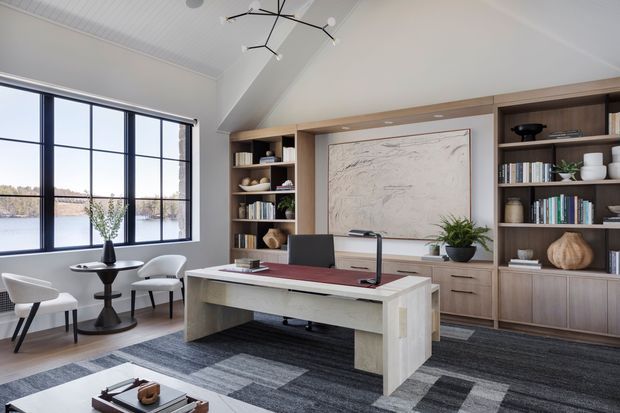 A refined home office with a large central desk, integrated timber shelving and cabinetry, a wall-mounted artwork panel, and a pair of occasional chairs by expansive black-framed windows. The room has a high vaulted white ceiling, layered grey patterned carpet, warm wood joinery, and a contemporary sculptural ceiling light. A calm waterfront landscape is visible beyond the glazing.