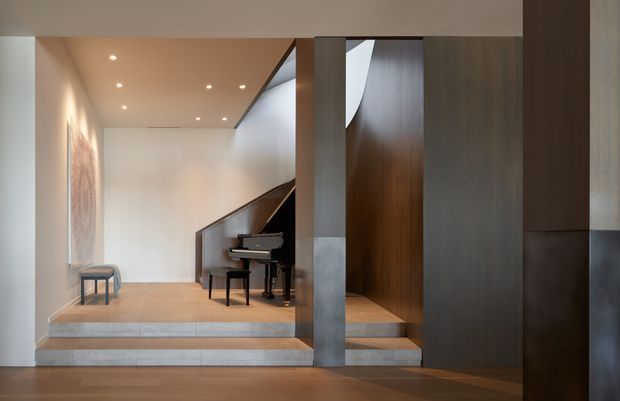 A refined contemporary interior featuring a small raised music platform with a grand piano, framed by dark timber wall panelling and a sculptural staircase or enclosed stair structure. Warm timber flooring, pale plaster walls, recessed ceiling lighting, and tiled step risers create a calm, gallery-like entry or circulation space.