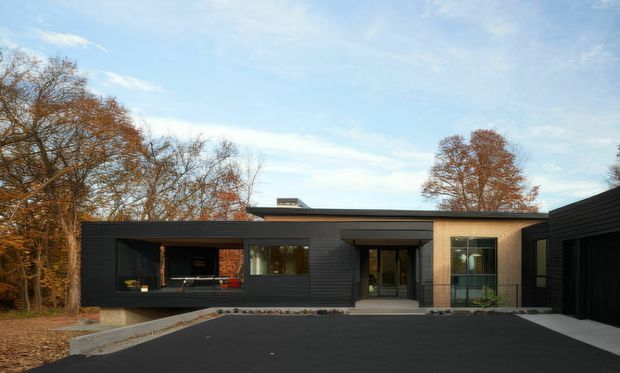 Single-storey contemporary residence with a low, flat roofline and strongly horizontal composition. The dark charcoal facade uses horizontal cladding, contrasted by a warm timber-toned entrance and adjoining light-toned wall section. Large glazed openings, a covered outdoor opening, and a paved dark driveway face a wooded suburban setting with mature deciduous trees.