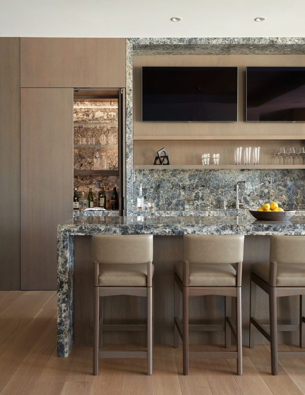 A refined contemporary kitchen and home bar area featuring full-height warm timber cabinetry, a stone-clad island and backsplash, integrated open shelving, two wall-mounted black screens, a concealed bottle display, and upholstered bar stools. The space has warm wood flooring, layered neutral tones, and dramatic blue-grey veined stone surfaces.