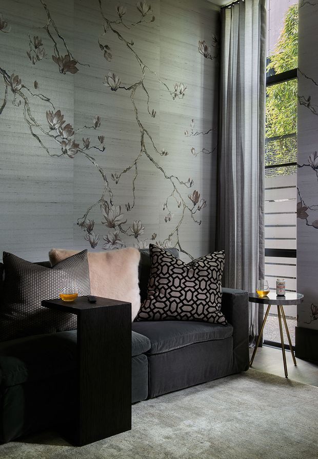 A refined, moody sitting area with two deep charcoal upholstered lounge chairs, patterned and textured cushions, and narrow black side tables. The walls are clad in a dramatic pale grey botanical mural or veined stone-like feature finish, complemented by full-height grey curtains and a tall black-framed window. A soft grey rug, small lamps, and warm drinkware create an intimate, luxurious atmosphere.