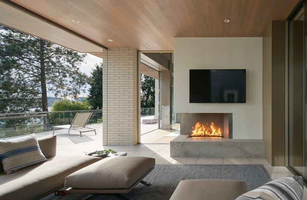 Contemporary open-plan living room with a large sectional sofa and ottoman facing a wall-mounted television and a sleek linear fireplace. The room opens onto a spacious outdoor terrace through extensive glazing, with a white brick structural column, warm timber-lined ceiling, polished light flooring, and a broad view of surrounding trees. The atmosphere is bright, refined, and inviting.