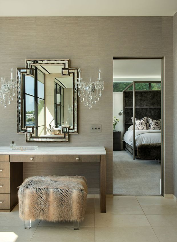 A refined dressing or bedroom vanity area with a long timber console featuring drawers, a pale stone top, an ornate studded framed mirror, and crystal wall sconces. A fur-covered stool sits in front. Through the open doorway, a bedroom is visible with a dark four-poster bed, patterned cushions, and a textured dark feature wall. Large pale floor tiles and softly textured taupe wallcovering create a luxurious, neutral interior.