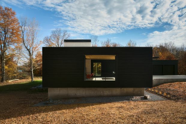 A low, rectilinear single-storey modern house set within a wooded site. The dark horizontal-clad facade is raised on a raw concrete base, with a centrally positioned timber-framed window and a small flat-roofed volume visible behind. Autumn trees and leaf-covered ground surround the building.