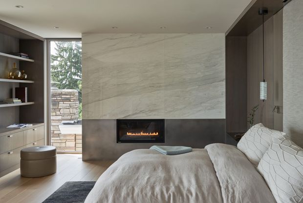 A refined contemporary bedroom with a low-profile bed facing a built-in linear fireplace. The room features a large pale veined stone feature wall above a dark hearth surround, dark timber built-in shelving and cabinetry, recessed ceiling lighting, a suspended pendant, and a glazed opening onto a stone outdoor area. The restrained neutral palette and layered textiles create a calm, luxurious atmosphere.