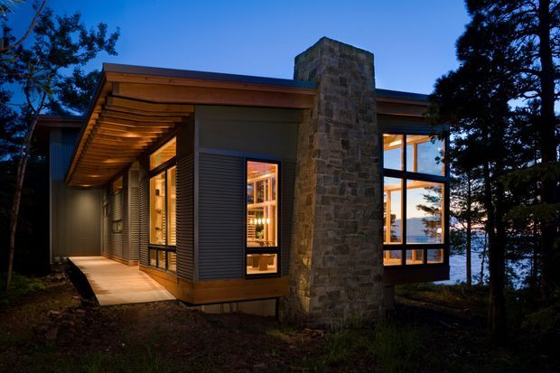 A modern, single-storey house set among trees beside visible water, photographed at dusk. The building features a low-slope roof with a broad timber-lined overhang, extensive dark-framed glazing, dark horizontal cladding, warm interior lighting, and a prominent rough natural-stone chimney rising through the roof. A timber-decked entry walkway leads along the side of the house.
