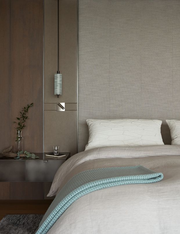 A sophisticated bedroom detail with a neatly made bed, pale textured bedding, white quilted pillows, and a folded pale blue throw. The room features a dark timber built-in wall and low cabinetry, taupe upholstered or textured wall panels, integrated bedside lighting, and restrained decorative styling with a small vase of greenery. The overall look is calm, warm, and refined.