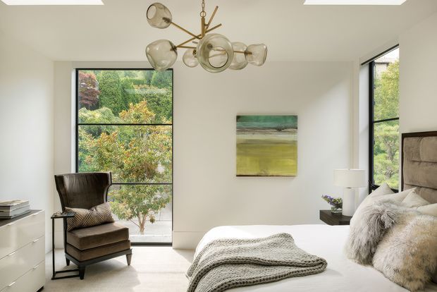 Bright contemporary bedroom with a large upholstered bed, layered neutral bedding, a textured throw, and a sculptural lounge chair beside expansive black-framed windows. White walls and cabinetry create a clean backdrop, while a brass-and-glass statement pendant, abstract artwork, and views of leafy landscaping add warmth and visual interest.