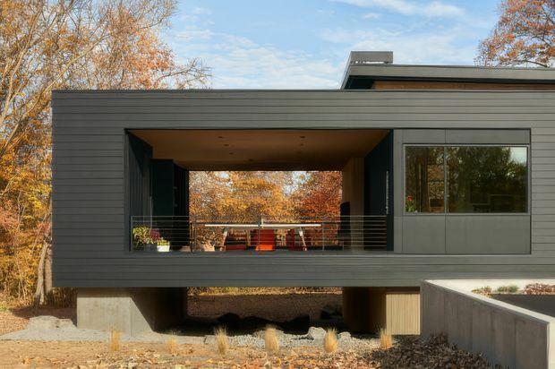 Exterior view of a contemporary, elevated flat-roof house set among mature deciduous trees. The dark horizontal-clad facade frames a deep recessed balcony or covered outdoor room with dining furniture, while large windows and an open lower level emphasize the modern structure and connection to the surrounding landscape.