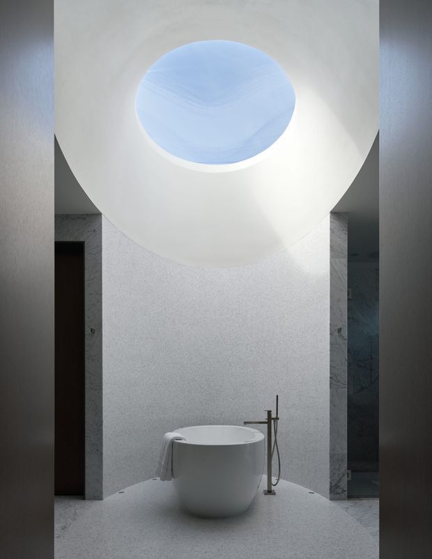 A serene minimalist bathroom centered on a freestanding white oval bathtub beneath a large circular skylight or ceiling opening. The room features pale textured walls and flooring, dark slender bath fixtures, a towel draped over the tub, and a partially visible glass-enclosed shower area at right. Soft daylight creates a calm, sculptural atmosphere.