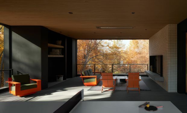 A contemporary open-plan living room opening onto a terrace or balcony, with expansive glazing overlooking autumn trees. The room features dark built-in joinery, a warm timber ceiling, a dark floor, a white tiled fireplace wall, and striking orange and green lounge chairs around a dining or entertaining area. A large central island or counter occupies the foreground.