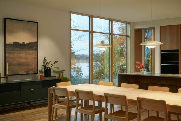 A warm, refined dining room with a long light-wood dining table and matching wooden chairs, positioned beside expansive timber-framed windows overlooking trees and water. A dark built-in sideboard, framed artwork, plants, pendant lights, and a dark kitchen or storage joinery zone complete the space.