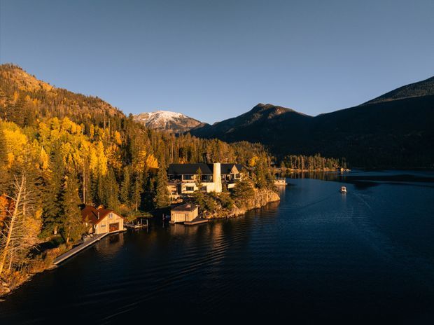 A scenic aerial or elevated view of a lakeside residential property surrounded by dense forest and mountainous terrain. The main house and smaller waterfront structures sit on a wooded shoreline beside a dark blue lake, with golden autumn foliage and snow-dusted peaks in the background.