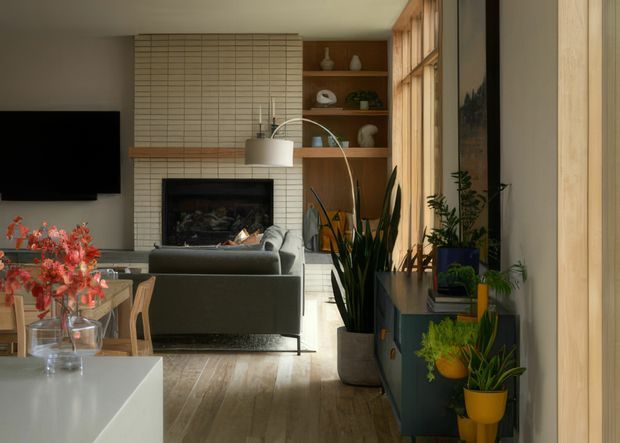 Warm, contemporary living room viewed toward a tiled fireplace wall, with a wall-mounted television, built-in timber shelving, large timber-framed glazing, layered indoor plants, and a compact dining area in the foreground. The room combines neutral finishes, timber flooring, dark cabinetry, and abundant natural light.