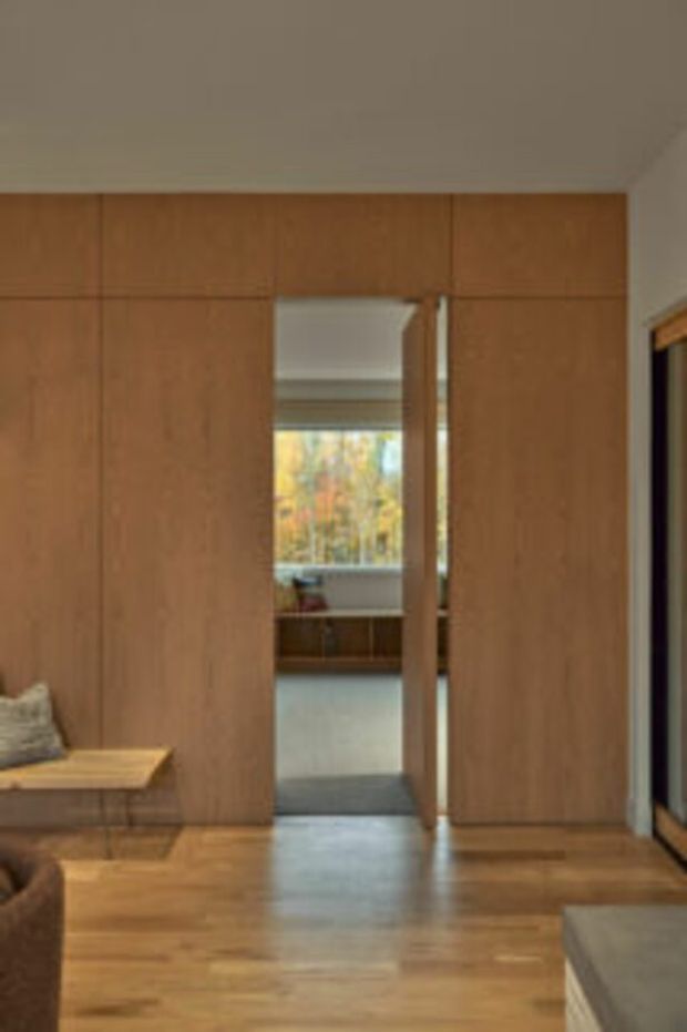 Warm interior view dominated by full-height timber wall panelling and integrated timber joinery, with a narrow doorway opening toward a bright adjoining room and window. Timber flooring, a built-in bench with cushion, and partial upholstered furniture are visible. The design is understated and natural, with clean linear detailing and warm wood tones.
