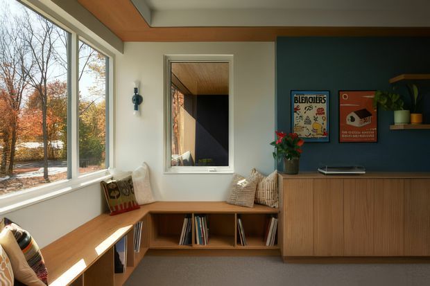 Warm mid-century-inspired living room or lounge corner with extensive corner glazing overlooking trees, built-in timber seating and shelving, a low-profile timber sideboard, and a deep blue feature wall. The room has a timber-lined ceiling with exposed beams, neutral walls, textured cushions, framed artwork, and abundant natural light.
