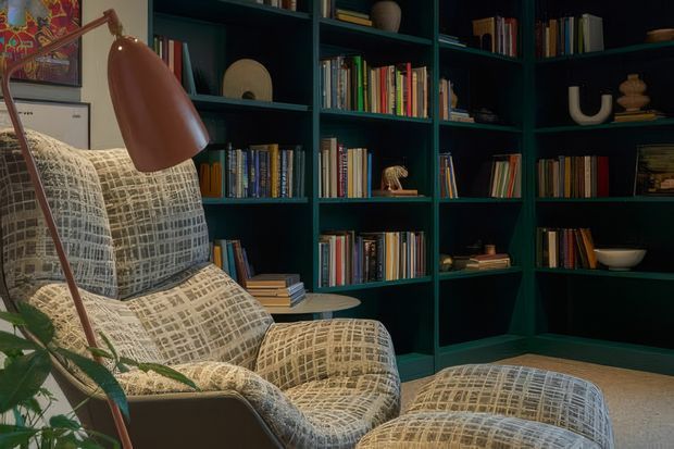 A moody, richly layered reading corner featuring a patterned upholstered lounge chair and matching ottoman, a slim copper-toned floor lamp, and extensive dark teal built-in shelving filled with books and curated objects. The space has a cosy, sophisticated mid-century-inspired character with warm timber flooring and low, atmospheric lighting.